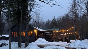 Eine Hütte mit einem mit Schnee bedeckten Deck, umgeben von Bäumen, mit Lichterketten.