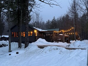 Eine Hütte mit einem mit Schnee bedeckten Deck, umgeben von Bäumen, mit Lichterketten.