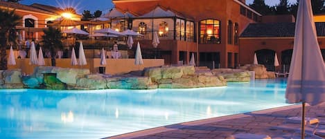 A swimming pool with lounge chairs and umbrellas, surrounded by a building with multiple levels and outdoor seating.