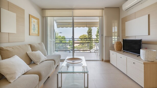 A modern living room with a sofa, a glass coffee table, a TV on a cabinet, and a balcony with a view.