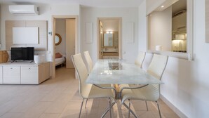 A modern living room with a flat-screen TV, a dining table with chairs, and a kitchen area with a large window.