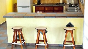 Cozinha compacta com parede amarela, geladeira, micro-ondas e três banquetas de madeira.