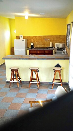 Een compacte keuken met een gele wand, een koelkast, een magnetron en drie houten krukken.