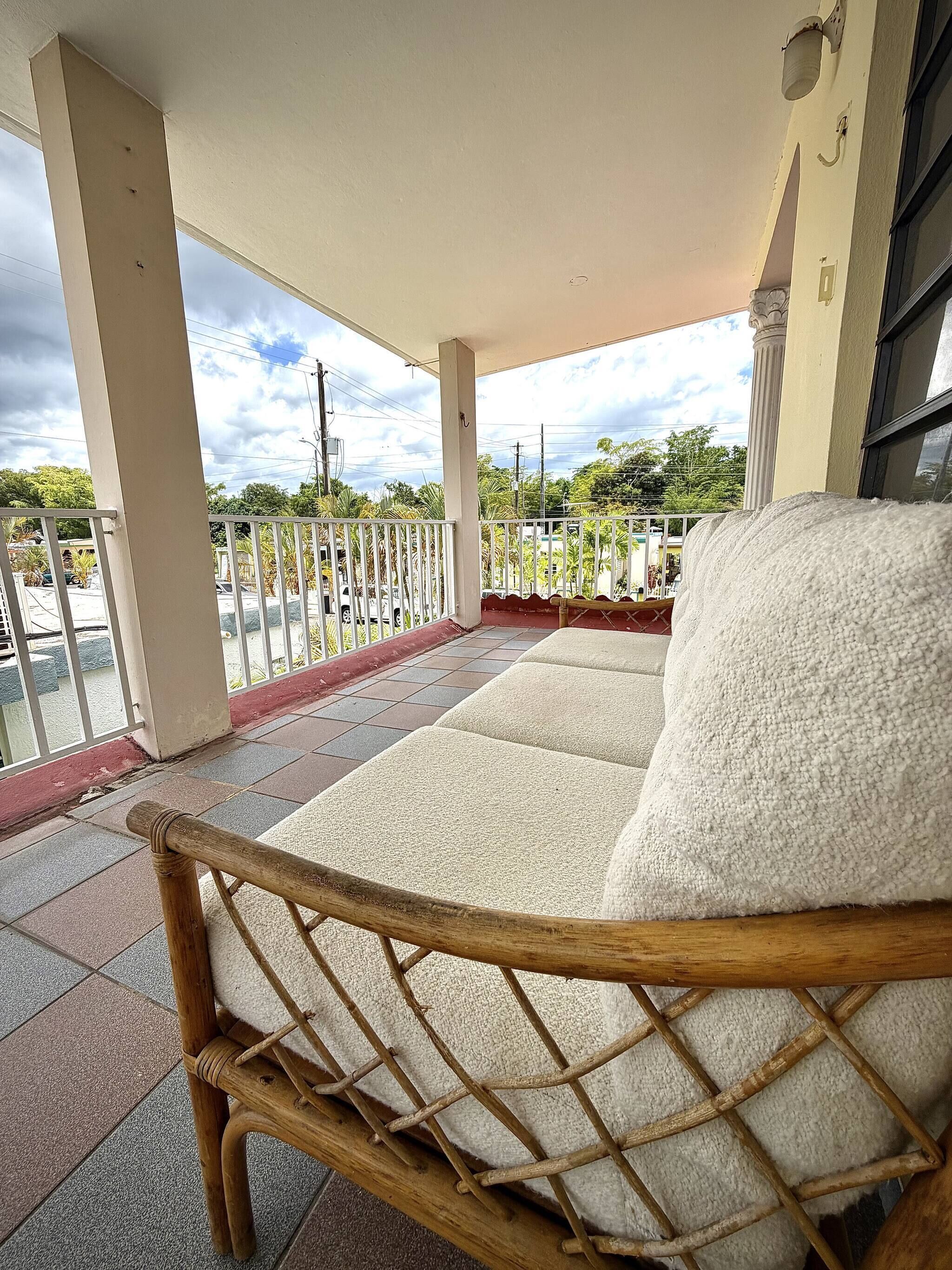 A covered outdoor seating area with a wooden chair, a white cushioned sofa, and a metal railing.