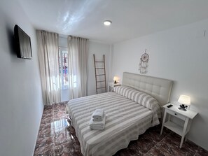 A bedroom with a bed, two bedside tables, a wall-mounted TV, a dreamcatcher, a window with curtains, and a view of the outside.
