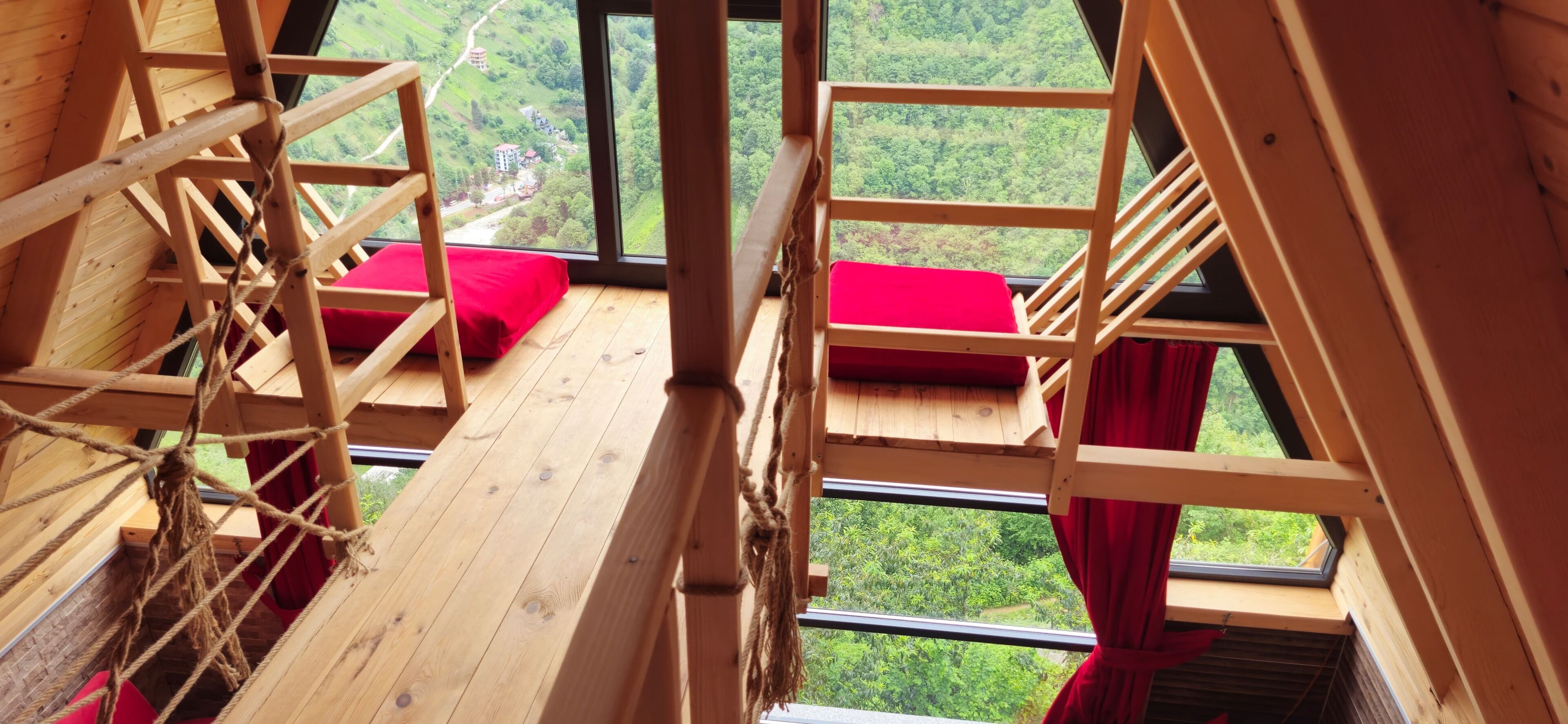 A wooden room with a view of a green landscape, a rope ladder, and red cushions.