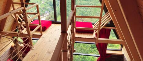 A wooden room with a view of a green landscape, a rope ladder, and red cushions.