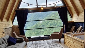 A high-ceilinged wooden interior with a sofa, chairs, a small table, and a view of a mountainous landscape through large windows. - HAVALI BUNGALOWS (Camlihemsin)