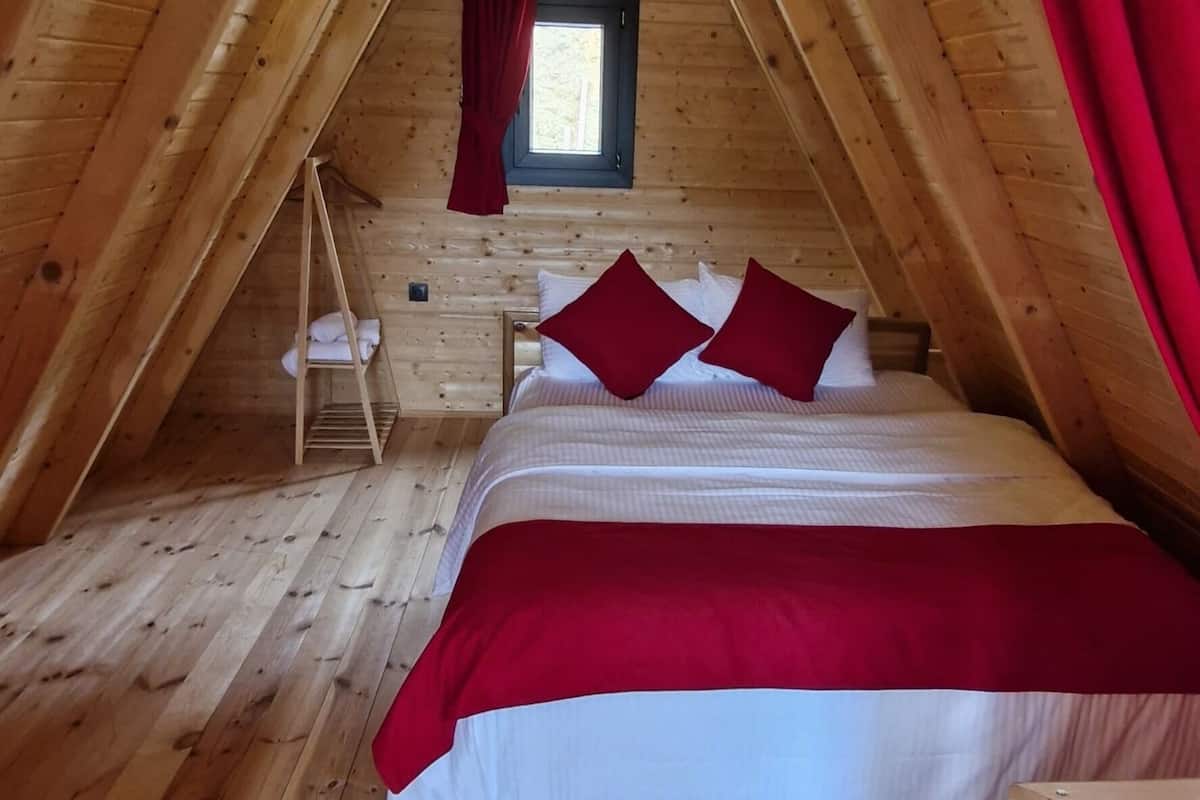 An attic bedroom with a wooden floor, a bed with red bedding, a window with curtains, and a ladder.