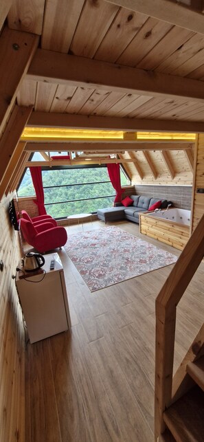 Un acogedor dormitorio en el ático con techo de madera, un sofá, una mesita con hervidor y vistas a un bosque a través de amplias ventanas.
