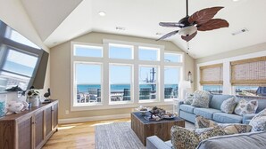 A spacious living room with a large window overlooking the ocean, a ceiling fan, and a cozy seating area. - High Tide Hideaway | Lux Ocean View in Kitty Hawk (Kitty Hawk)