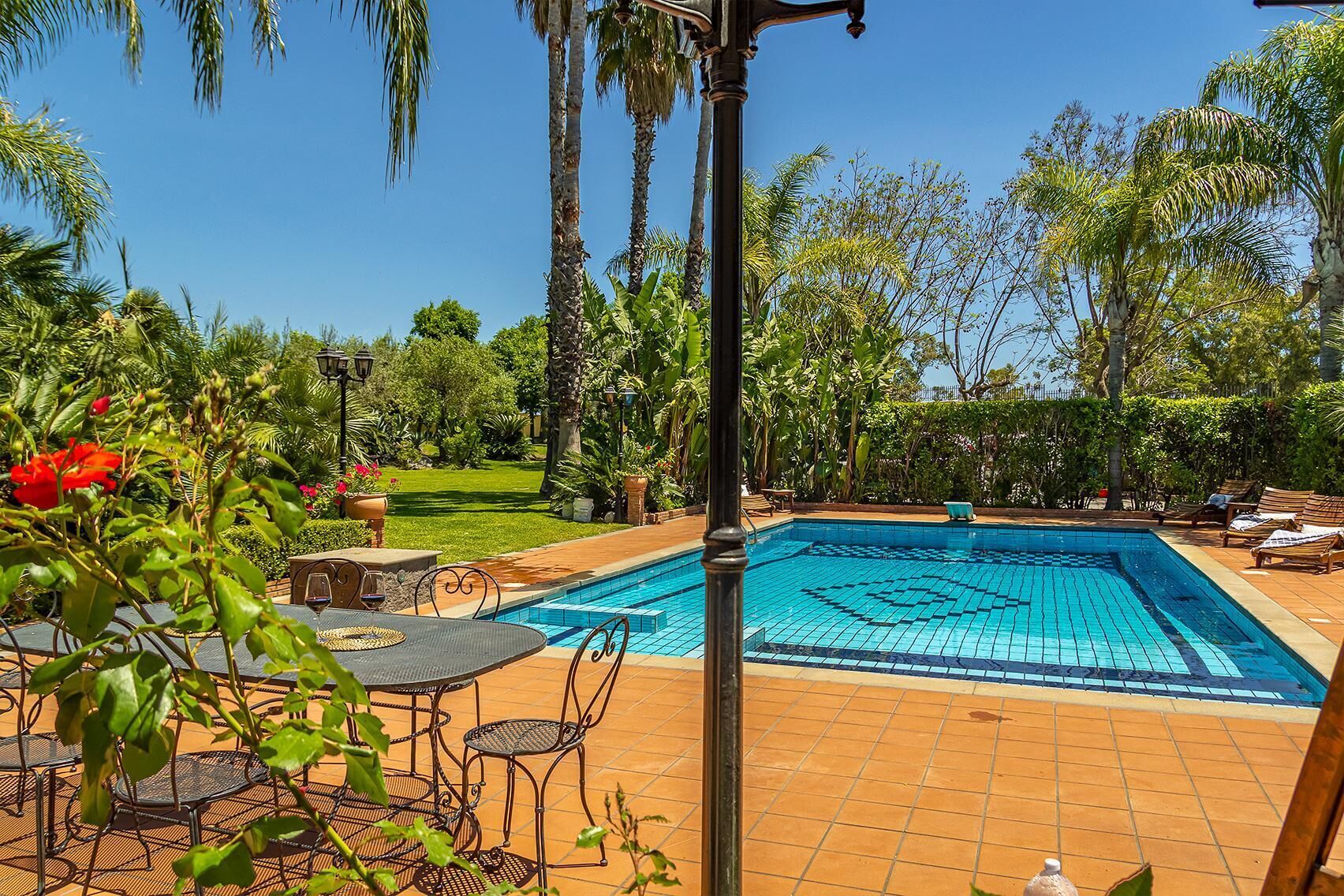 A rectangular swimming pool with a tiled deck, surrounded by palm trees and a well-maintained lawn.