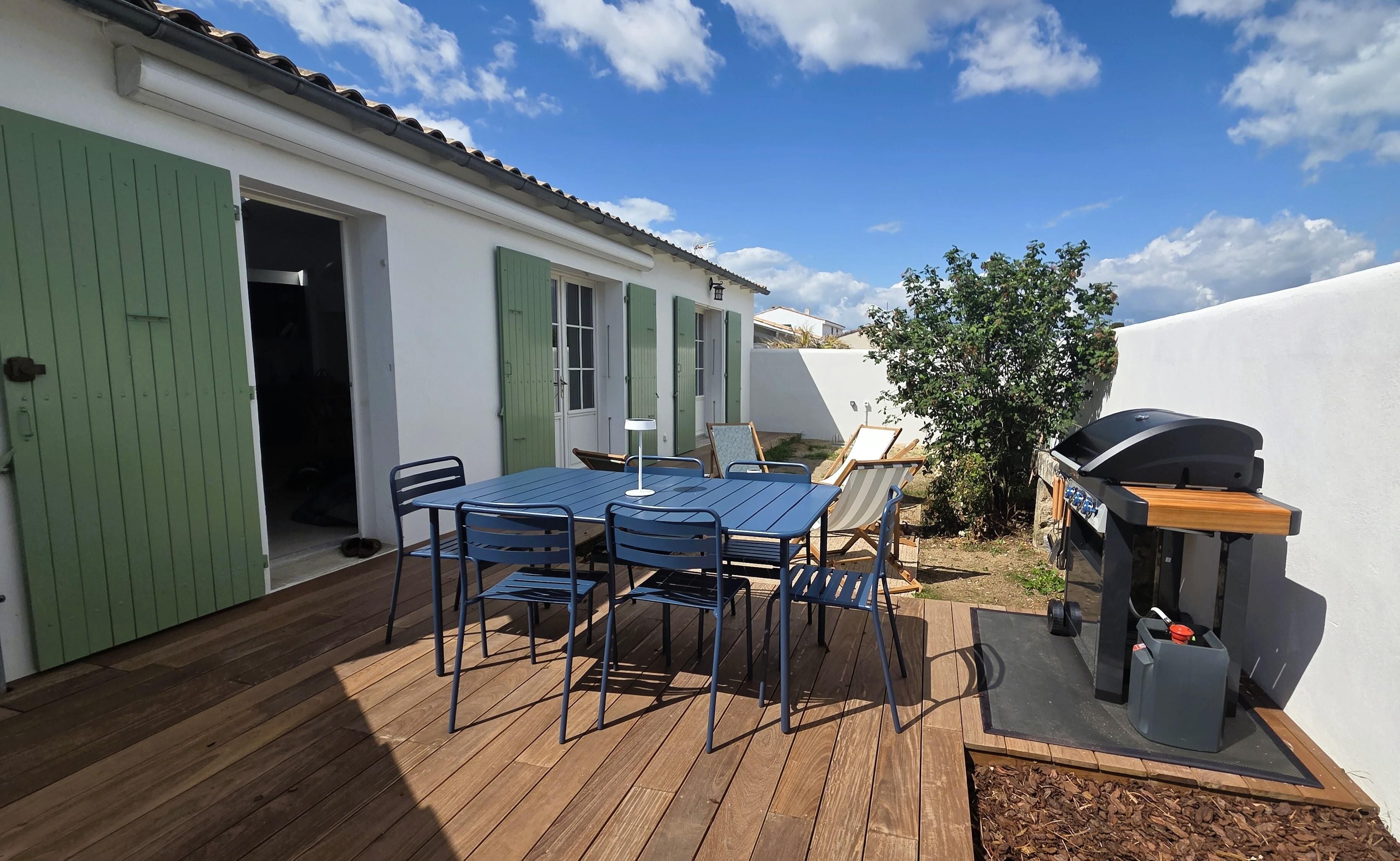 Une terrasse avec une table et des chaises, un barbecue et une porte avec des volets verts.