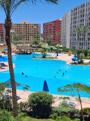 A large swimming pool surrounded by palm trees and blue umbrellas, with buildings in the background. - lagoons and golf Arya view (Marsa Matrouh Governorate)