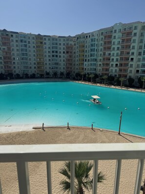 Una piscina grande y clara de color azul, rodeada por una playa de arena, con un edificio de varios pisos al fondo.