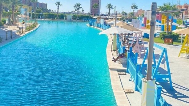 A swimming pool with a blue water feature, surrounded by a deck with umbrellas and lounge chairs. - lagoons and golf Arya view (Marsa Matrouh Governorate)