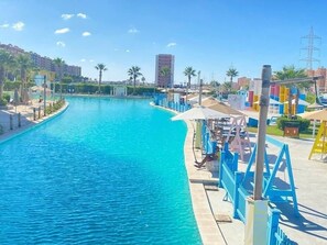 Una piscina con un elemento acuático azul, rodeada por una terraza con sombrillas y sillas reclinables.