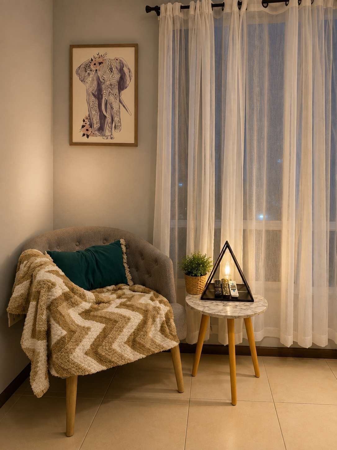 A cozy room with a sofa, a patterned throw blanket, a small side table with a plant and a lamp, and a framed picture on the wall.
