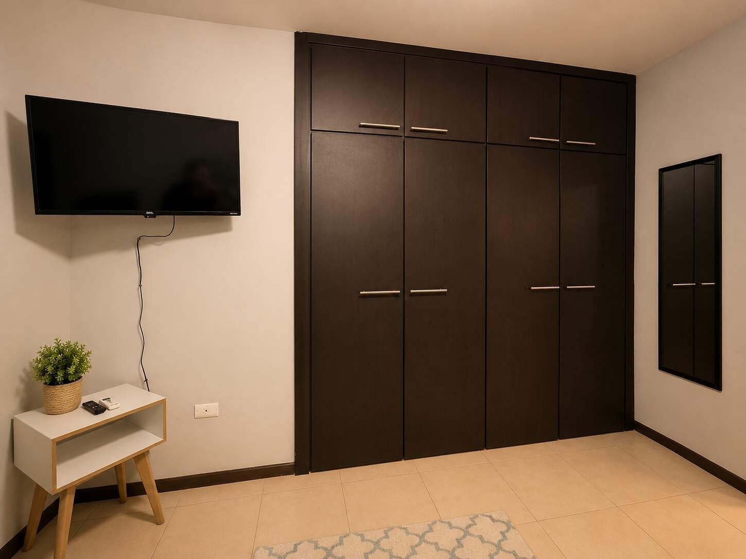 Une pièce avec une armoire de couleur sombre, un téléviseur à écran plat fixé au mur, une petite table console blanche ornée d’une plante, et un tapis à motifs sur le sol.