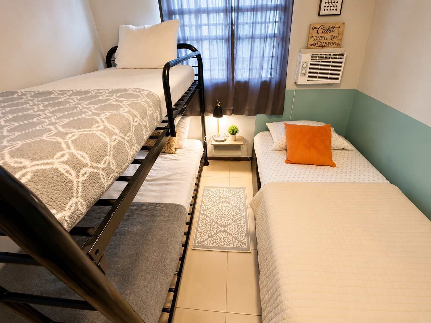 A room with two beds, a small table with a lamp, a potted plant, and a wall sign.