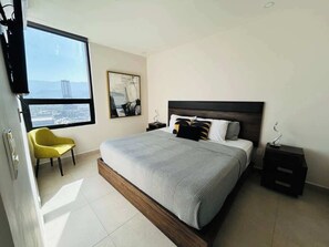 A modern bedroom with a large bed, a yellow chair, a nightstand, and a view of the city through a window.