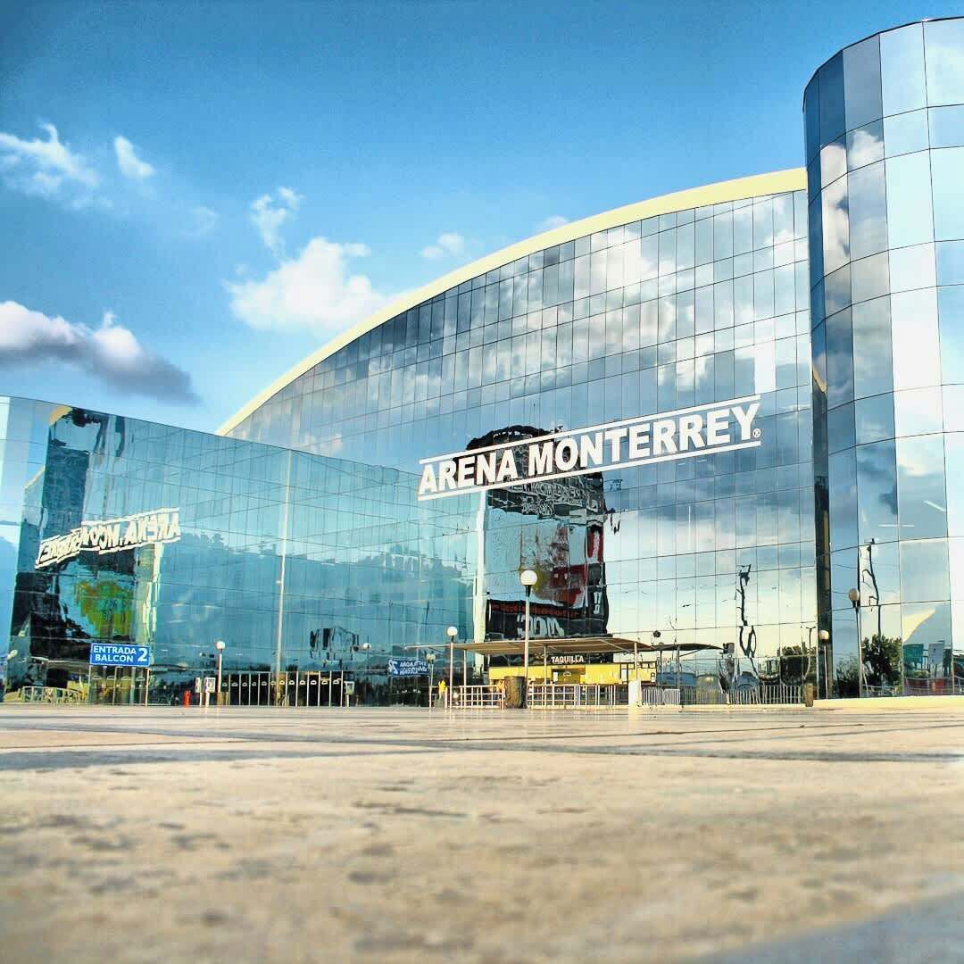 A modern glass building with 'ARENA MONTERREY' signage, a clear sky, and a paved area in front.