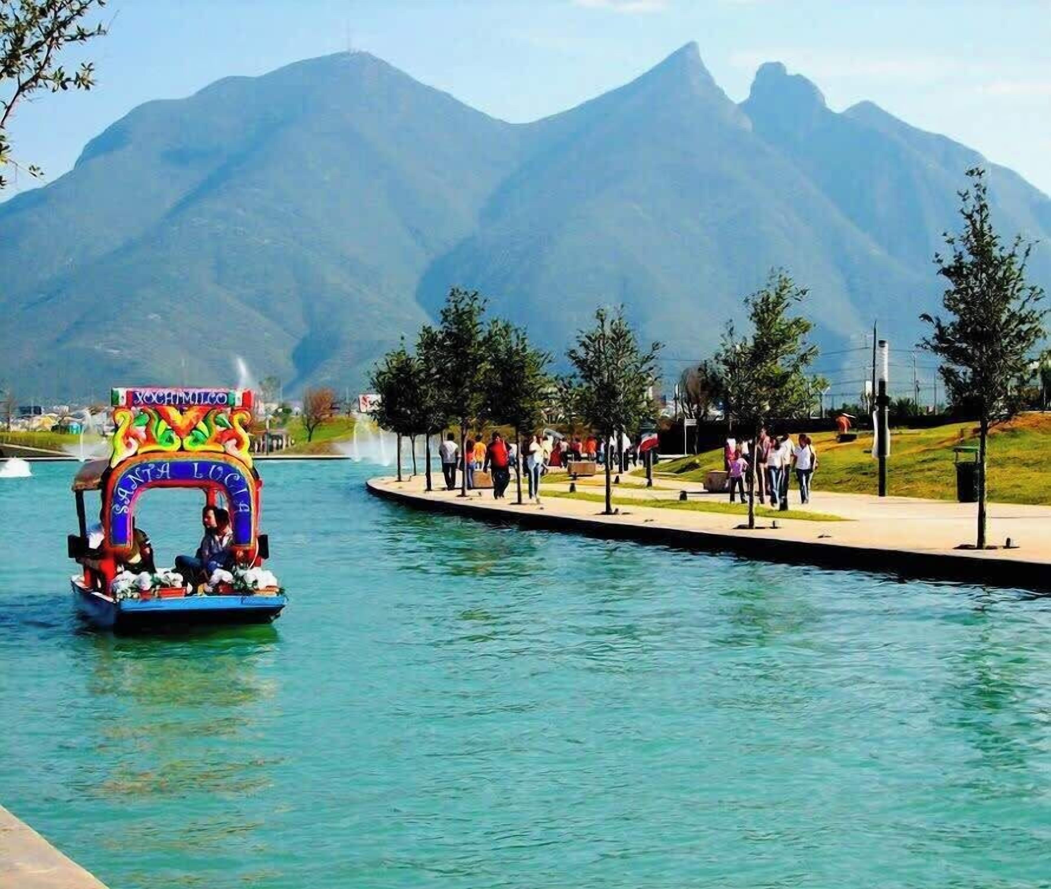 A boat with a colorful archway labeled 'Xochimilco' and 'Santa Lucia' on a calm waterway with people walking along the shore and mountains in the background.