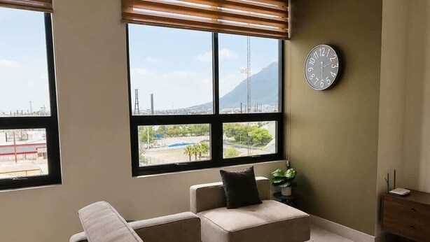 A living room with a sofa, a small table, a clock on the wall, and large windows with a view of a cityscape. - Fundidora Loft w/Parking & AC, Near Arena (Monterrey)