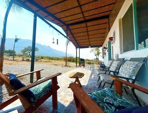 A covered outdoor seating area with wooden furniture and patterned cushions, a small table, and a view of distant mountains. - Hermosa Estancia Para Familia/10px (Urrutias)