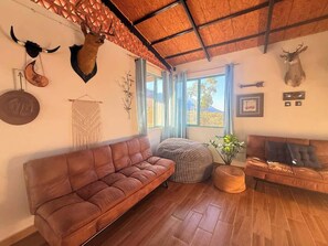 A cozy living room with a brown leather sofa, a round woven ottoman, and a wooden floor. - Hermosa Estancia Para Familia/10px (Urrutias)