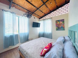 A bedroom with a wooden ceiling, a bed with patterned bedding, a red pillow, a TV on the wall, and a window with sheer curtains. - Hermosa Estancia Para Familia/10px (Urrutias)