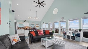 Ett rymligt vardagsrum med en svart lädersoffa, ett soffbord i glas och en grå pall. Längre in i rummet finns ett köksområde med utsikt över havet genom stora fönster.
