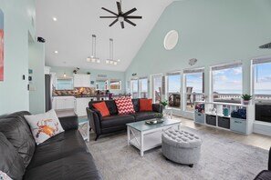 A spacious living room with a black leather sofa, a glass coffee table, and a grey ottoman. There is a kitchen area in the background with a view of the sea through large windows. - Beachfront with Hot Tub & Putt Putt | Sleeps 16 (Gilchrist)