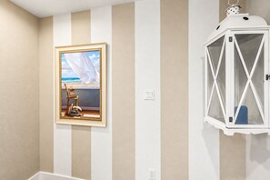 A room with a framed painting of a beach scene, a white lantern, and a chair with books. - Bolivar Beachfront | 6BR | Sleeps 28 | Spacious Deck & BBQ (Port Bolivar)