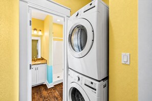 Un cuarto de lavado con dos lavadoras apiladas, una puerta que da a un baño con espejo, e interruptor de luz en la pared.