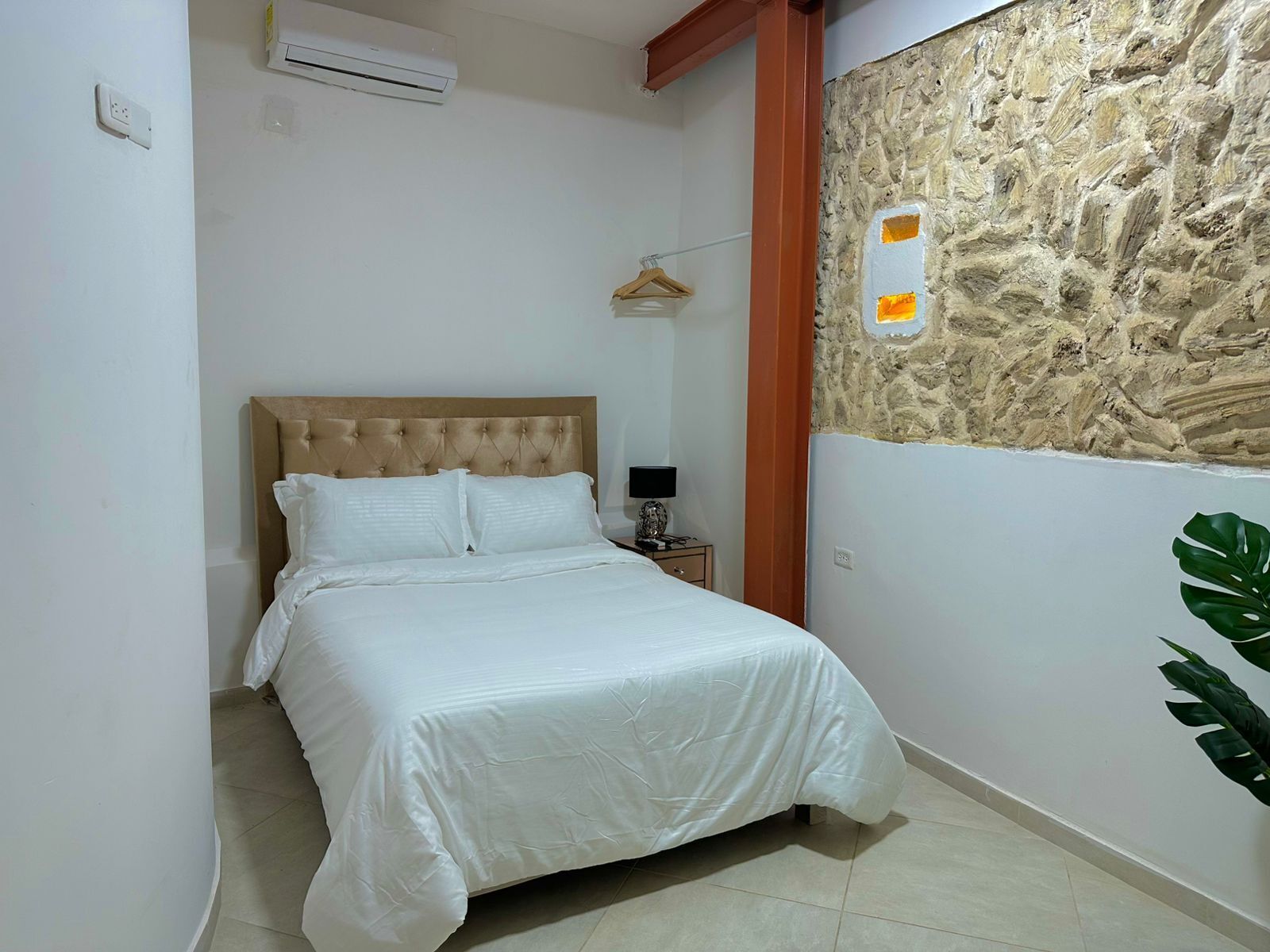 A bedroom with a bed, a nightstand, a wall-mounted air conditioner, and a stone wall with a small window.