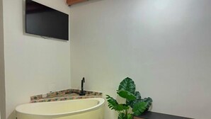 Ein Badezimmer mit Badewanne, Waschbecken, Spiegel, Zimmerpflanze und einem an der Wand befestigten Flachbildfernseher.