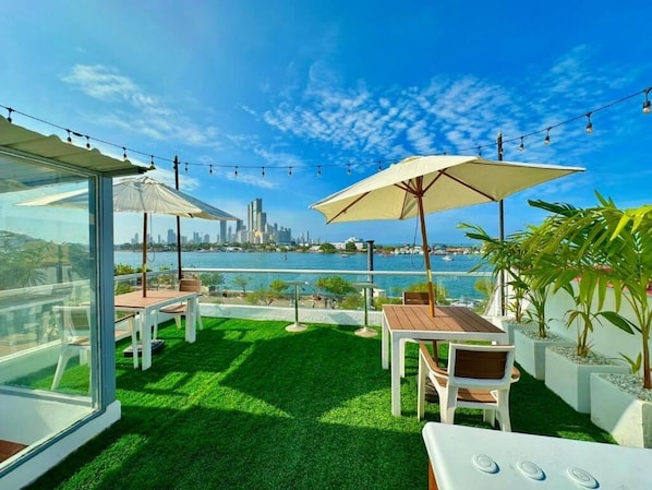 A rooftop terrace with a view of a city skyline, outdoor seating, and string lights. - Hotel Victoria Caps (Cartagena)