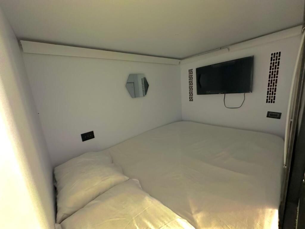 A small, single-bed room with a flat-screen TV mounted on the wall, a window air conditioner, and a power outlet.