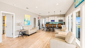 A spacious living area with a wooden floor, a dining table with chairs, a sofa, and a kitchen in the background. - Beachfront 5BR | Gulf Views + Game Room with Pool Table (Port Bolivar)