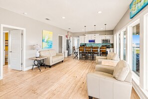 A spacious living area with a wooden floor, a dining table with chairs, a sofa, and a kitchen in the background. - Beachfront 5BR | Gulf Views + Game Room with Pool Table (Port Bolivar)