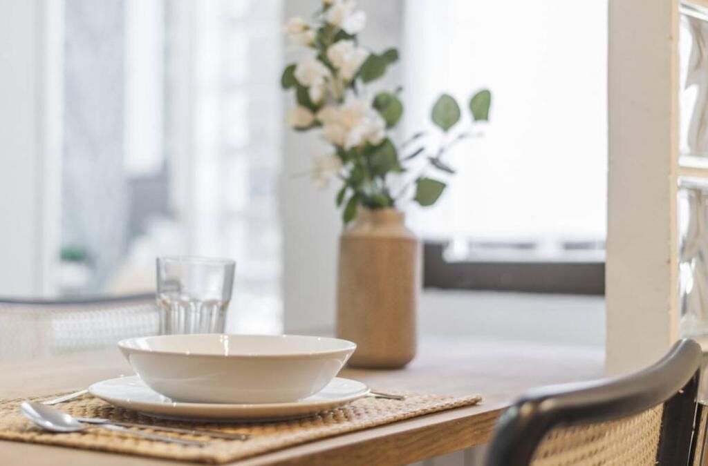 Ein Esstisch mit einer Schüssel, einem Glas und einem Löffel, einer Vase mit Blumen und einem Stuhl.