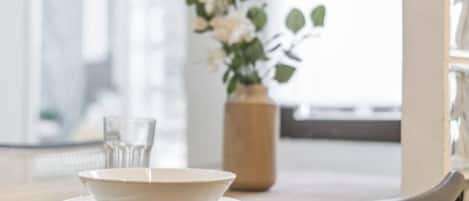 Una mesa de comedor con un bol, un vaso y una cuchara, un florero con flores y una silla.