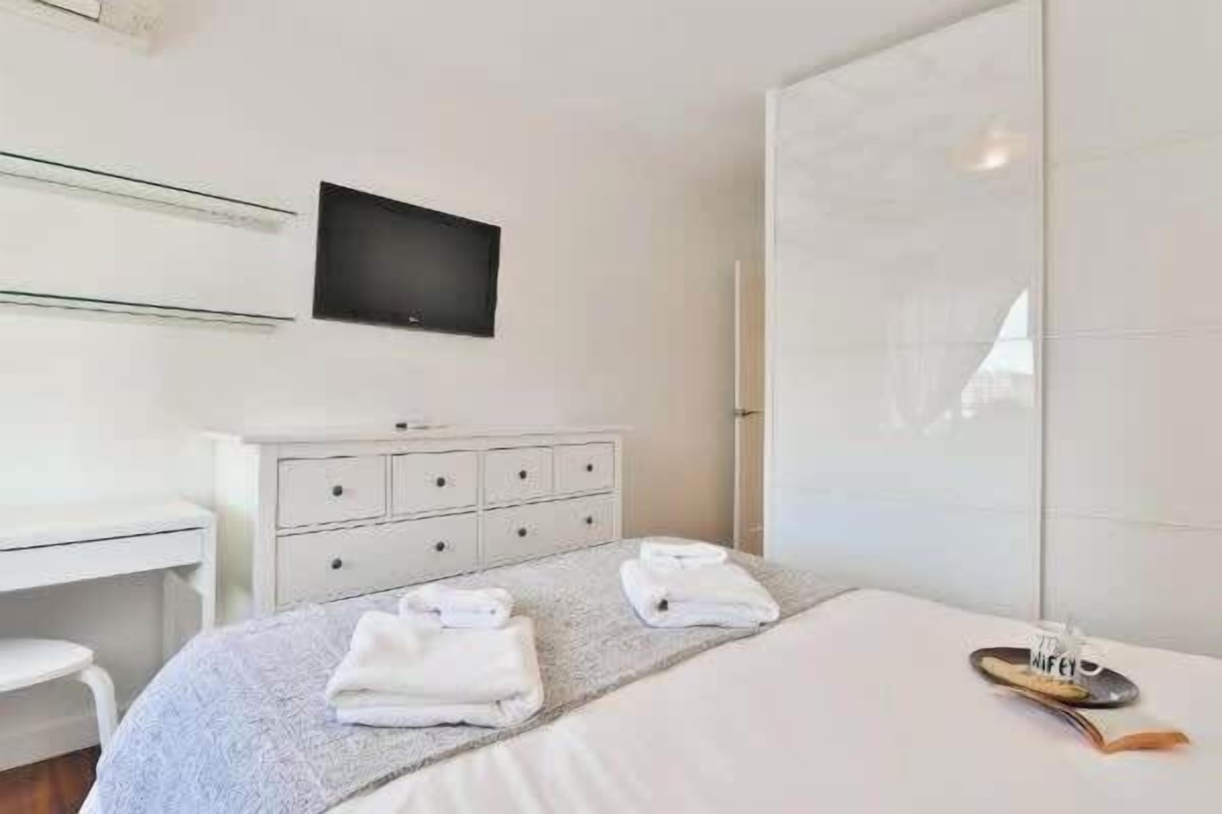 Ein Hotelzimmer mit Bett, einem weißen Schminktisch, einem Fernseher an der Wand und einem Paar Schuhe auf dem Bett.