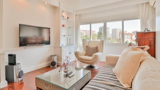 En moderne stue med et fladskærms-tv, et glasbord, en beige lænestol, en hvid sofa og et træskab.
