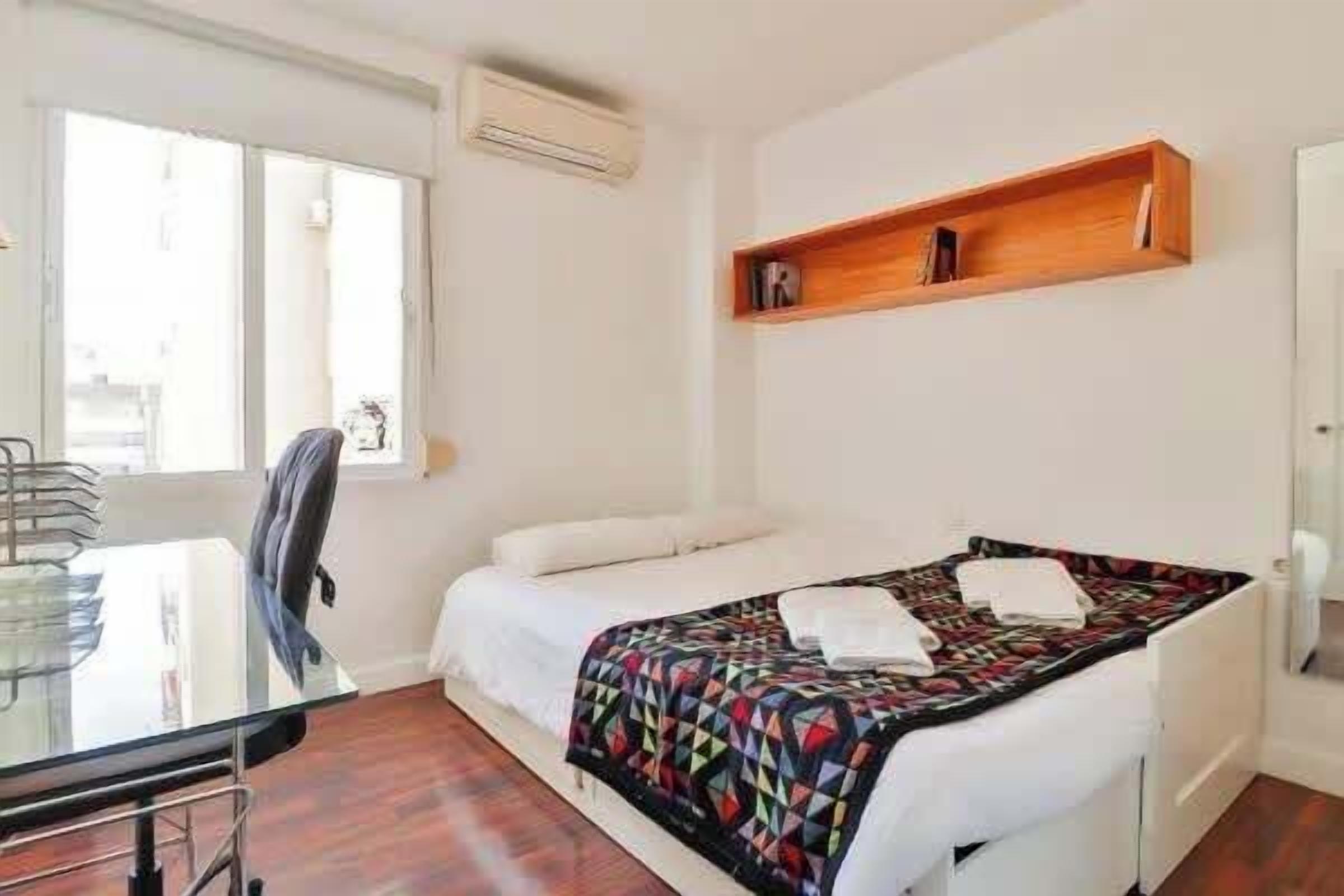 Une chambre d’hôtel avec un lit, un bureau, une chaise, une fenêtre et une étagère.