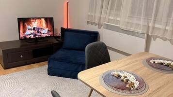 Ett rum med en tv-skärm som visar en öppen spis, en blå soffa, ett matbord med tallrikar och ett fönster med tunna, genomskinliga gardiner.