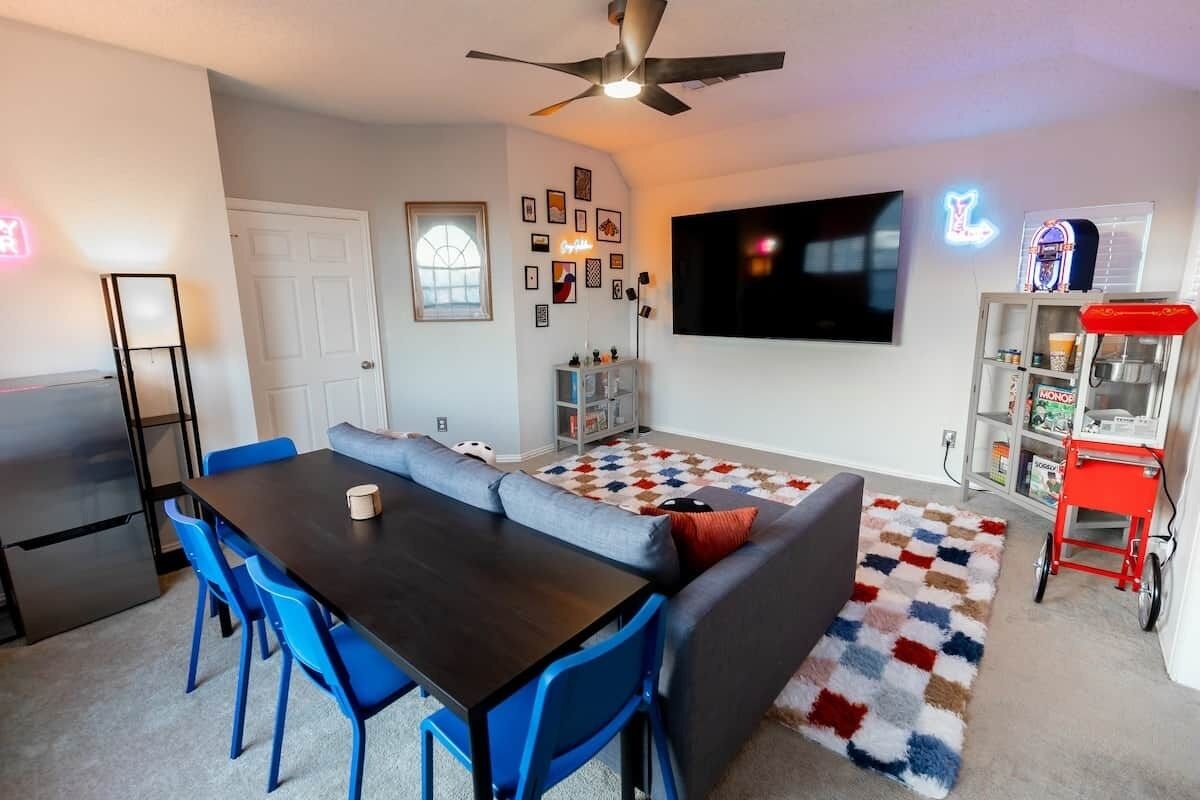 Un soggiorno moderno con un tavolo da pranzo in legno scuro, sedie blu, un divano grigio, una ventola a soffitto, un televisore a schermo piatto e un cartello luminoso al neon.