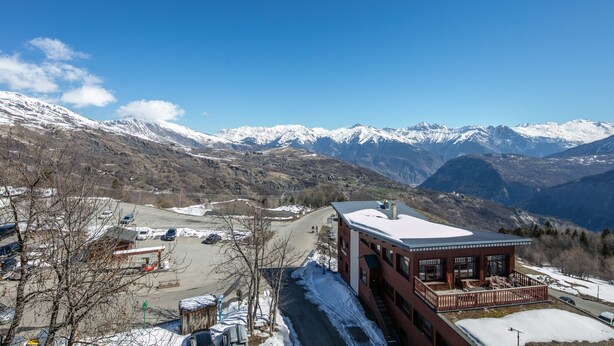 Eine Berglodge mit schneebedecktem Dach, einem Parkplatz und Blick auf verschneite Berge.