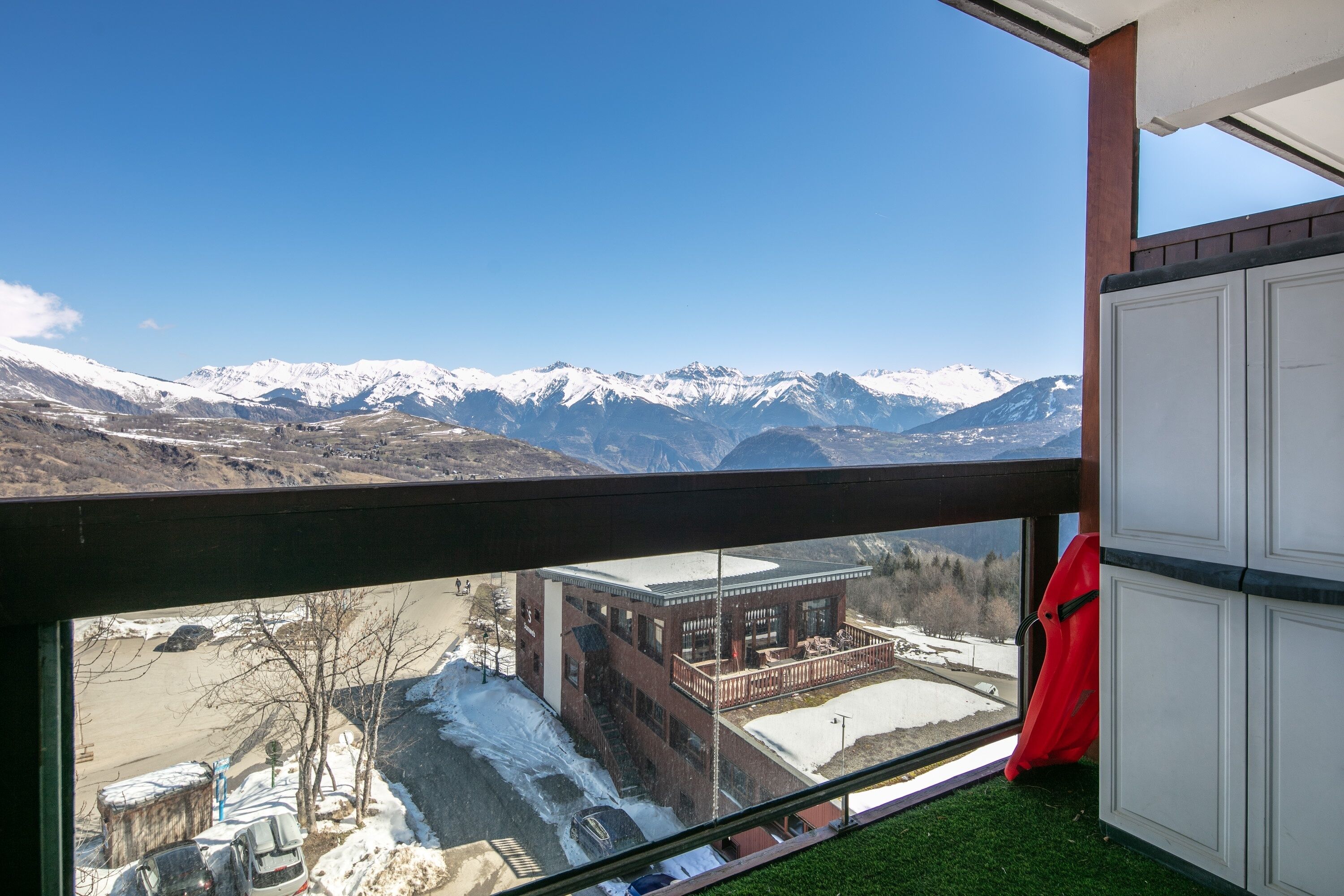 Una habitación con vistas a montañas nevadas, un balcón con una silla roja y un edificio de fachada de ladrillo.